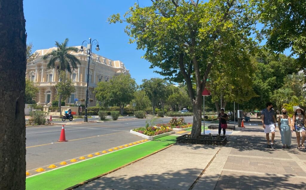 Pedestrians on the shaded sidewalk of Paseo de Montejo beside Palacio Cantón in Mérida